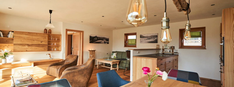 stadtalm toelzer land bad toelz urlaub in deutschland voralpenland in der natur in bayern in den bergen auf dem land im gruenen nachhaltige oekologische unterkunft holz lehm bienen lamas kubische bauweise alpinarchitektur ferienwohnung isarflimmern karwendelrauschen wohnbereich esstisch sofa barbereich kueche ledersessel